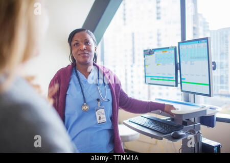 Female doctor looking at patient while standing by desktop computers in medical room Stock Photo