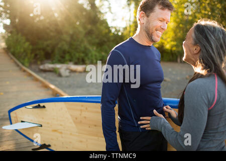 Cheerful couple with paddleboard looking at each other while standing on boardwalk Stock Photo