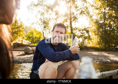 Smiling man holding water bottle while looking at woman sitting on pier Stock Photo