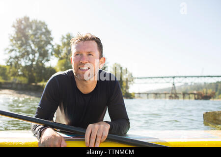 Man looking away while leaning over paddleboard on river against clear sky Stock Photo