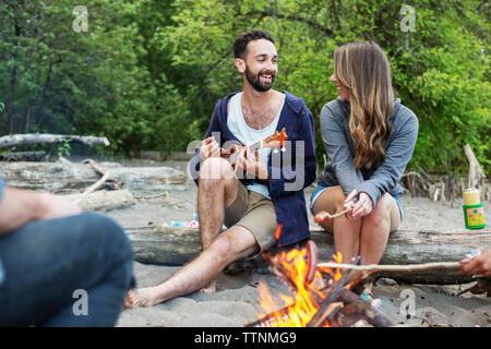 Man playing ukulele while sitting with friends by campfire at riverbank