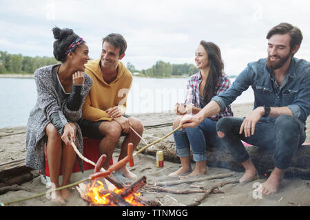 Happy friends roasting sausages while sitting at campfire against river Stock Photo
