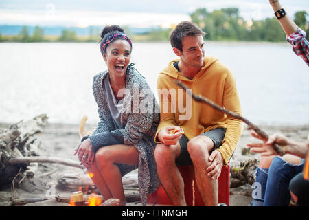 Friends sitting on cooler by campfire against river Stock Photo