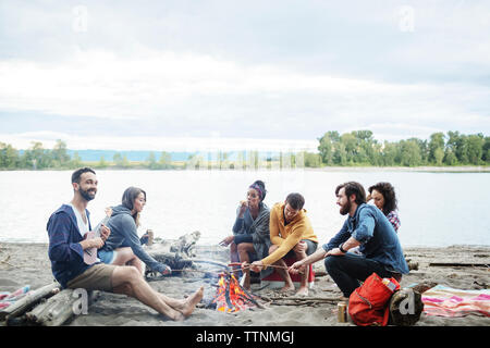 Happy friends roasting sausages on campfire at riverbank against sky Stock Photo