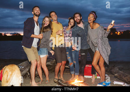 Happy friends at campfire by river against cloudy sky Stock Photo