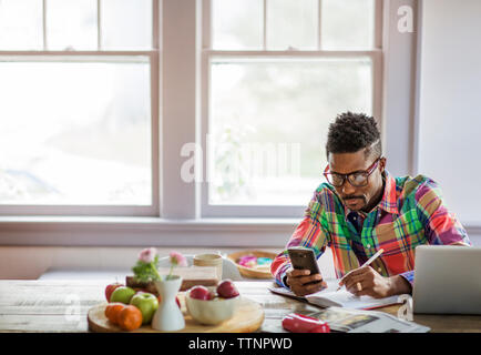 Man using smart phone while writing in book at home Stock Photo