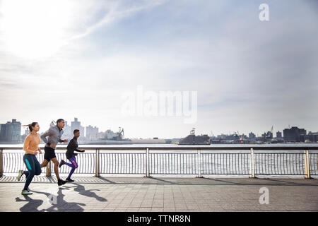 Side view of determined athletes running on footpath by river against sky Stock Photo