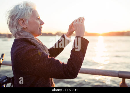 Senior woman photographing by sea during sunset Stock Photo