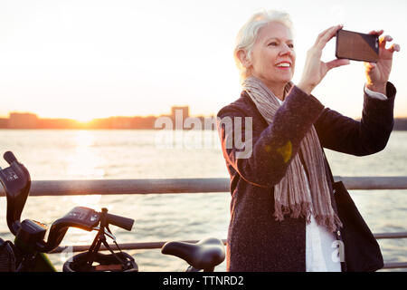 Happy senior woman photographing by sea during sunset Stock Photo