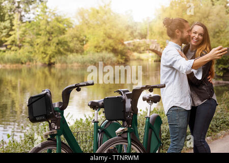 Man taking selfie while kissing girlfriend by lake Stock Photo