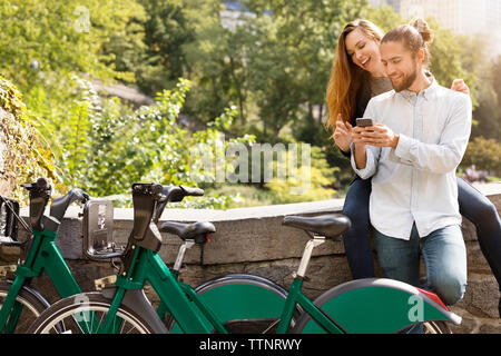 Couple using mobile phone while leaning on retaining wall Stock Photo