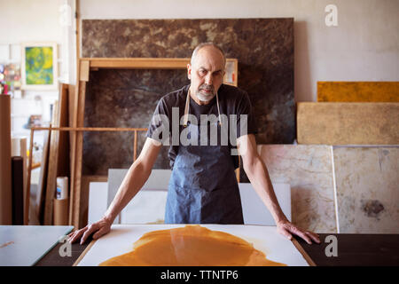 Portrait of senior artist standing by painting at table in workshop Stock Photo