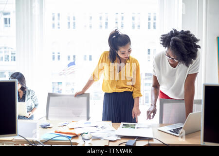businesswomen pointing towards document while standing against window with female colleague sitting in background Stock Photo