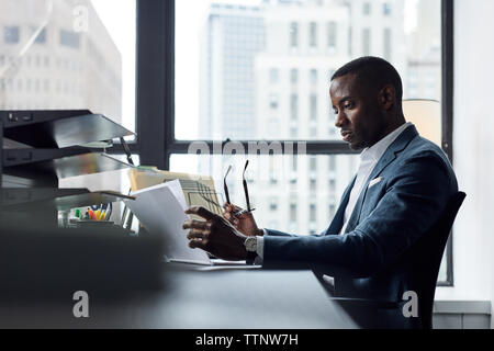Serious businessman reading documents while holding eyeglasses against window in office Stock Photo