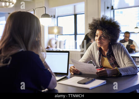 businesswomen discussing over documents with colleagues in background at creative office Stock Photo