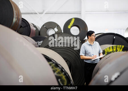 Manager writing on clipboard while analyzing rolled up metal sheets at Steel Mill Stock Photo