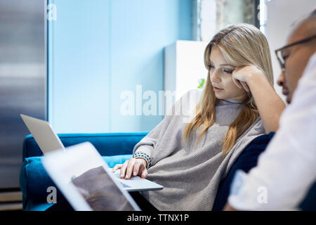 Male and female business colleagues using laptops while sitting on sofa at creative office Stock Photo