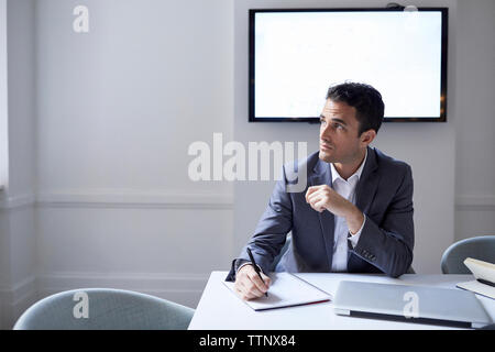Thoughtful businessman writing on spiral notebook while sitting at conference table Stock Photo