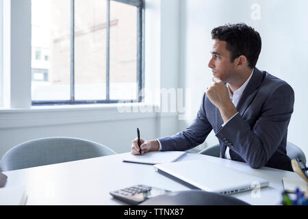 Thoughtful businessman writing on spiral notebook at conference table in board room Stock Photo