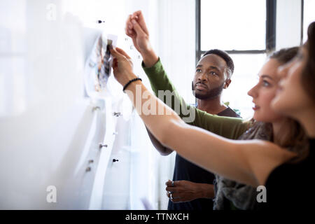 Serious businessman looking at female colleagues attaching photograph printouts on bulletin board in office Stock Photo