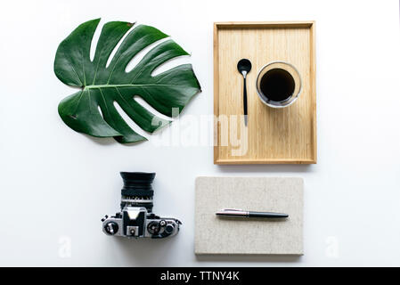 Overhead view of coffee in wooden tray with monstera leaf and camera by office supplies arranged on white background Stock Photo