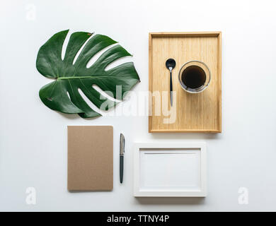 Overhead view of coffee in wooden tray with monstera leaf and picture frame by office supplies arranged on white background Stock Photo