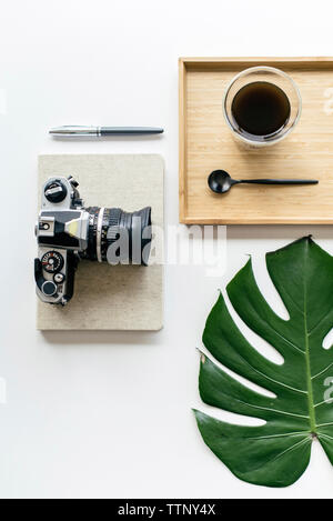 Overhead view of coffee in wooden tray with camera and office supplies by monstera leaf arranged on white background Stock Photo