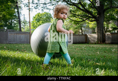 Rear view portrait of cute girl standing with fitness ball at lawn Stock Photo