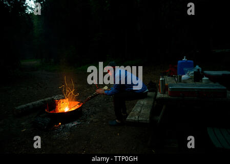 Side view of hiker warming hands by campfire at campsite during dusk Stock Photo