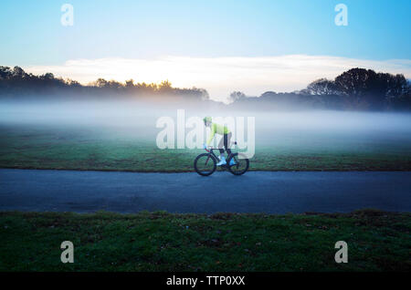 Side view of man cycling on street amidst grassy field in foggy weather