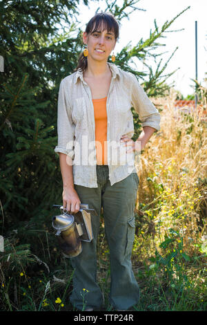 Portrait of confident beekeeper with hand on hip holding bee smoker while standing against plants at field Stock Photo