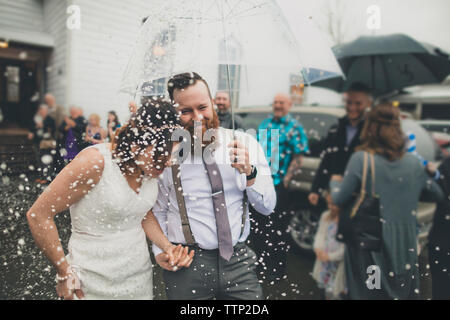 Happy newlywed couple walking with umbrella while people standing in background during wedding ceremony Stock Photo