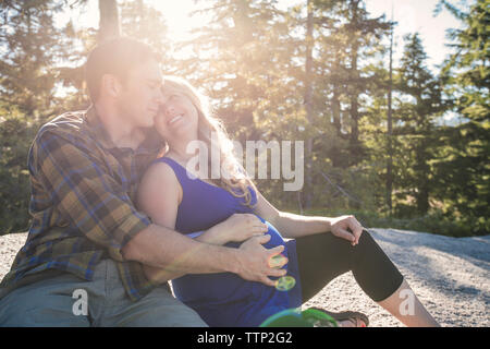 Man touching pregnant woman's stomach while sitting against trees during sunny day Stock Photo