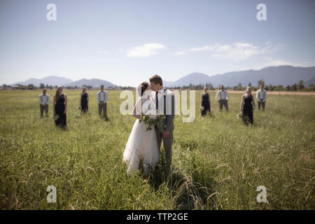 Newlywed couple kissing while standing at grassy field with groomsmen and bridesmaids in background during sunny day Stock Photo