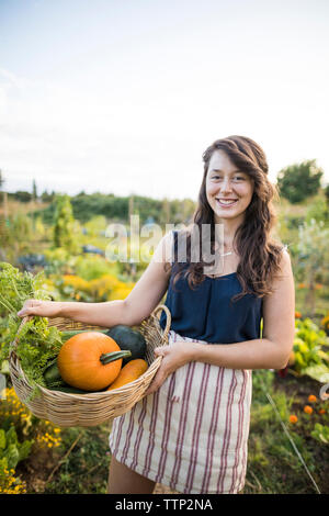Portrait of happy woman carrying vegetables in basket against clear sky at community garden Stock Photo