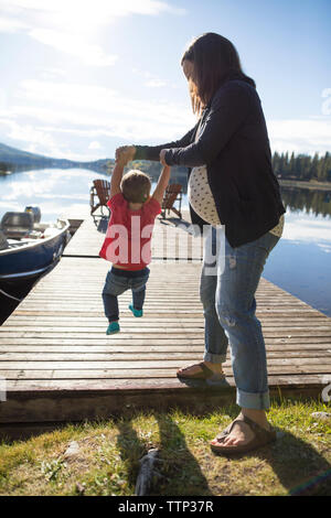 Full length of pregnant mother holding son's hands while swinging him on wooden pier by lake Stock Photo