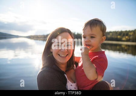 Portrait of cheerful mother carrying son against lake Stock Photo