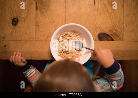 Overhead view of baby boy holding spoon in breakfast bowl on wooden table at home Stock Photo
