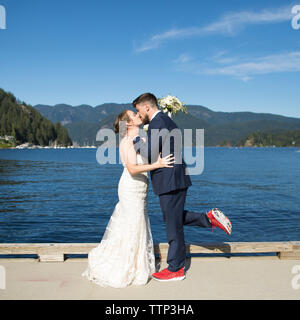 Side view of newlywed couple kissing while standing by lake against blue sky Stock Photo