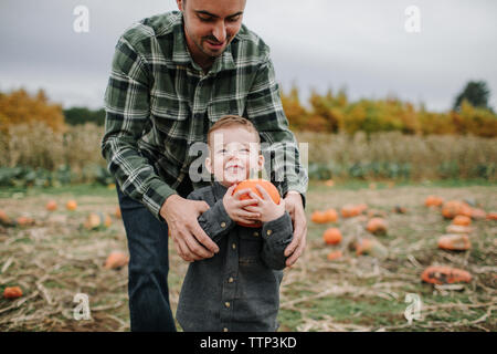 Father helping cute smiling son in holding pumpkin on field during autumn