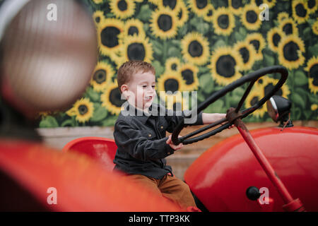 Cute smiling boy holding steering wheel while sitting on red tractor by sunflower farm Stock Photo