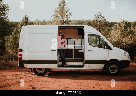 Man wearing cap and sunglasses while standing in motor home against sky at forest Stock Photo