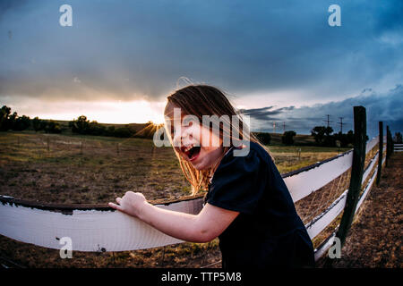Side view of happy cute girl laughing while standing by fence against cloudy sky during sunset Stock Photo