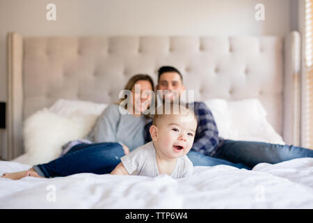 Baby boy laying on bed smiling with mom and dad in background indoors Stock Photo