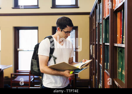 Confident man with backpack reading book while standing by shelf in library Stock Photo