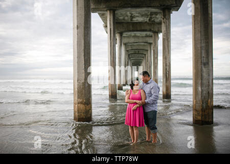 Smiling couple standing on shore under bridge at beach Stock Photo