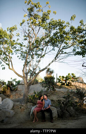 Couple talking while sitting on rock against sky in park during sunset Stock Photo
