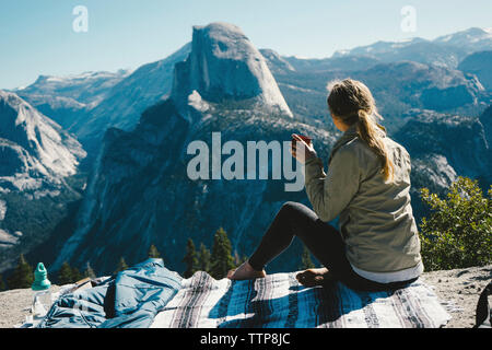 Full length of female hiker having drink while sitting on mountain at Yosemite National Park during sunny day Stock Photo