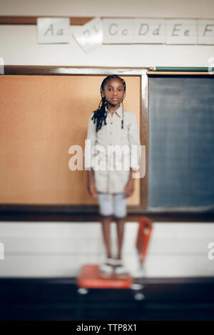 Portrait of girl standing on chair against blackboard in classroom Stock Photo