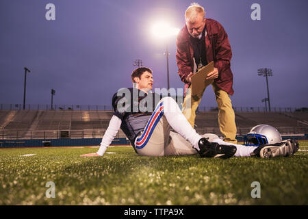 Coach showing clipboard to tired American football player relaxing on field at stadium Stock Photo